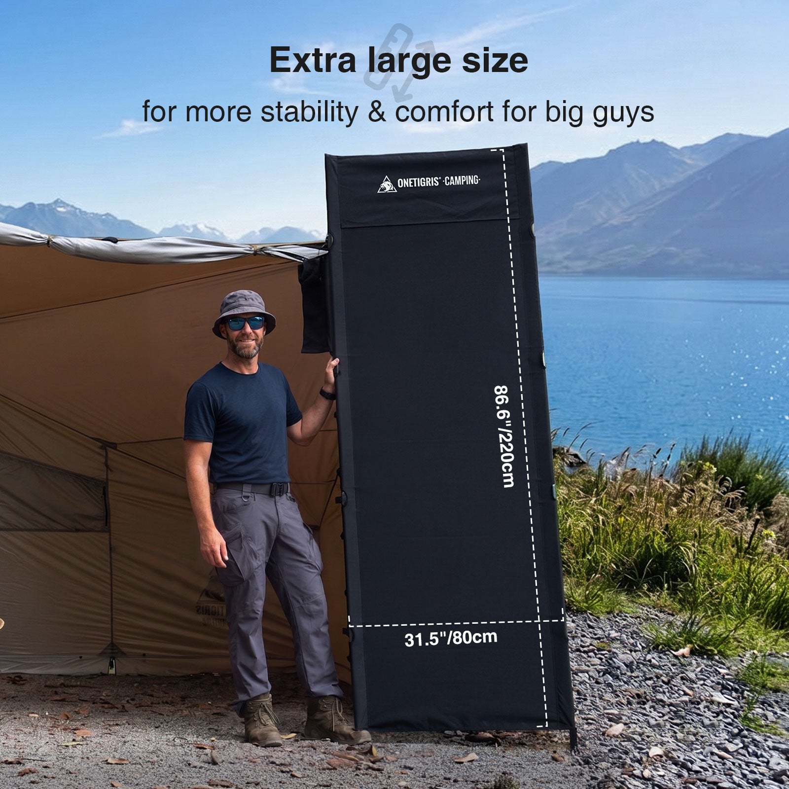 Person holding a large camping cot next to a tent with mountains and lake in the background