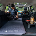 Person sitting on a camping mattress in the back of a van with cooking setup and 'Onetigris Camping' branding.