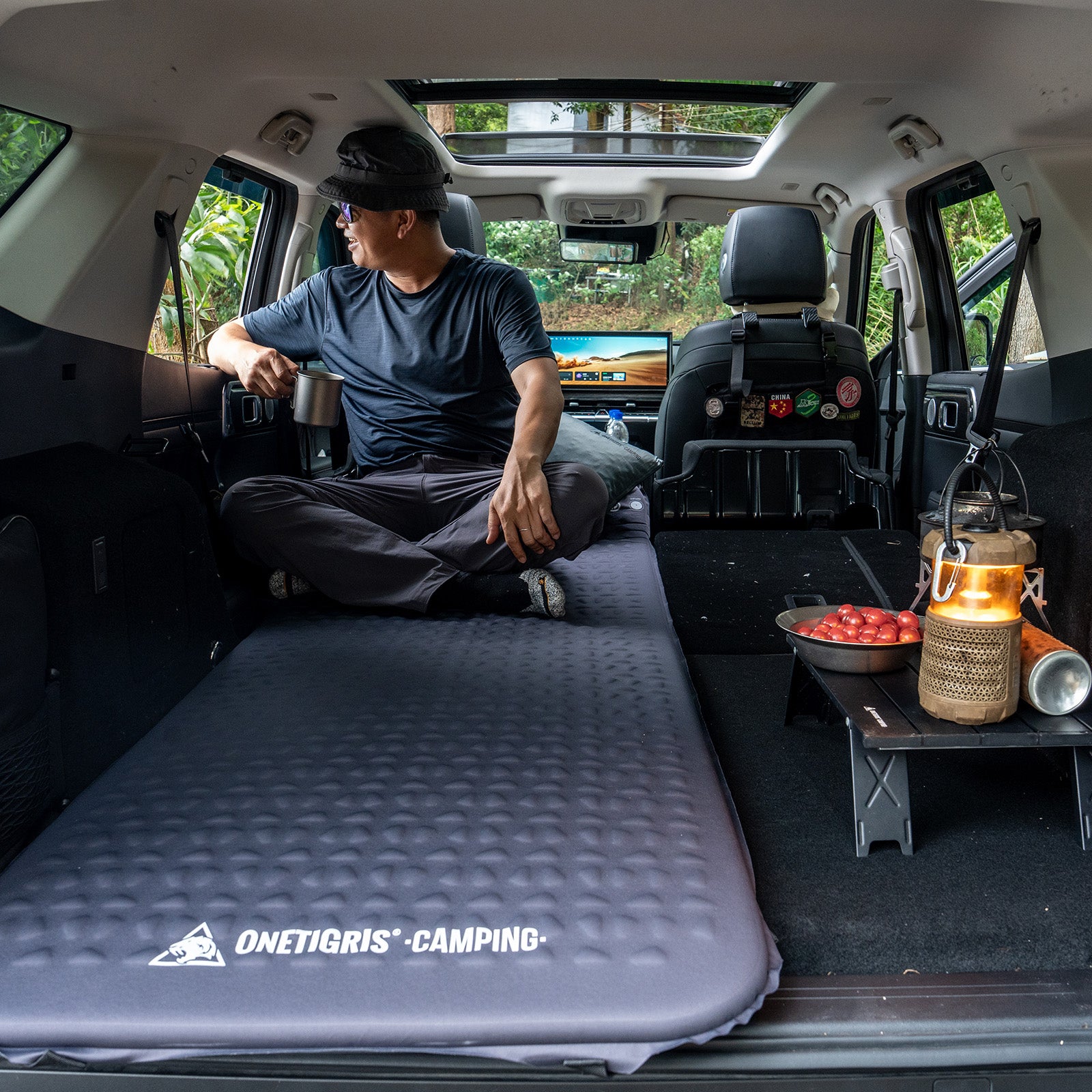 Person sitting on a camping mattress in the back of a van with cooking setup and 'Onetigris Camping' branding.