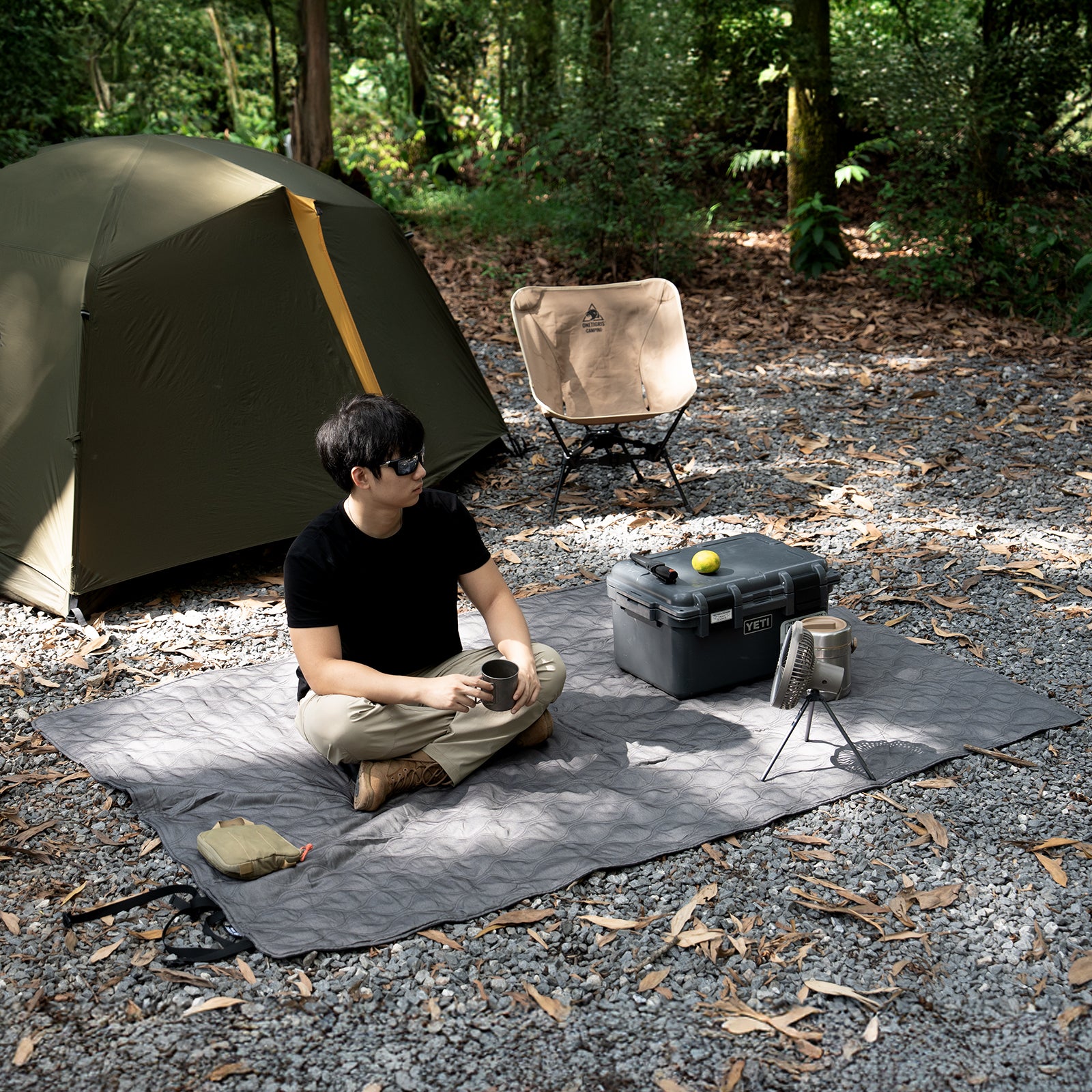 Person sitting on a camping mat with a tent and chair in a forest setting