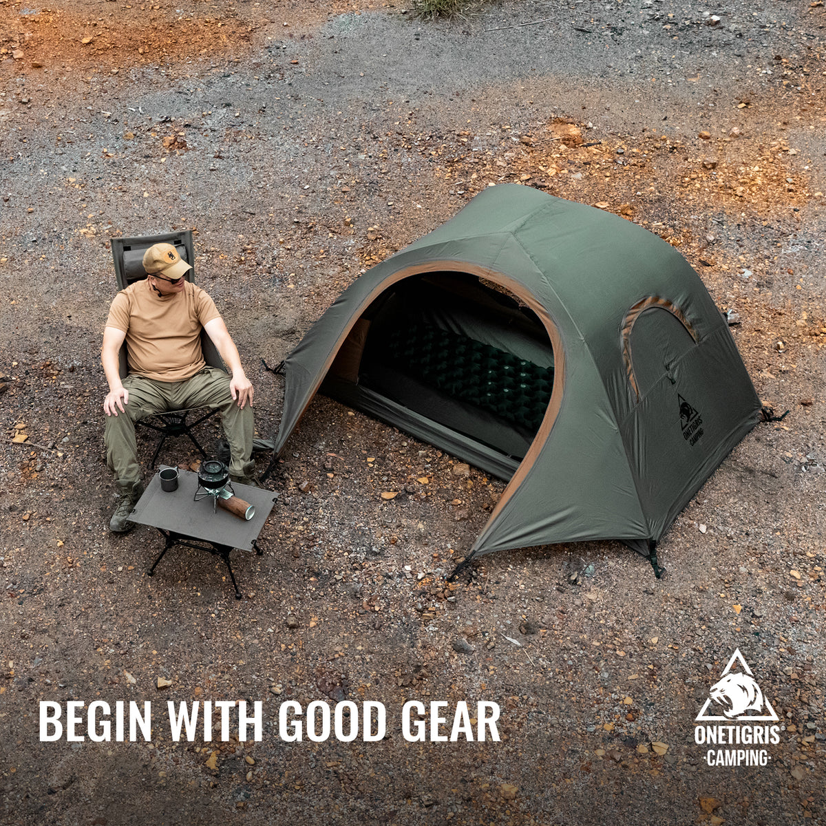 An overhead outdoor camping scene showing a man sitting on a folding chair beside an Onetigris BASTION  tent with a small folding table. The ground is gravel and dirt with scattered leaves.