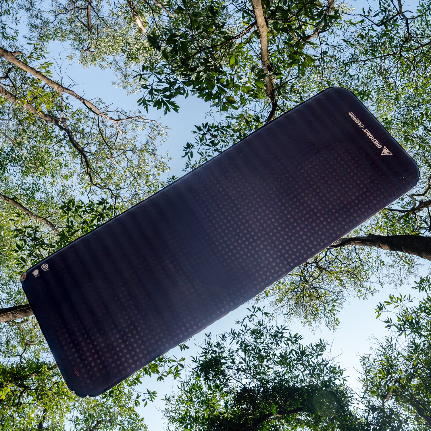 Black inflatable mat with brand logo against a natural background of trees and sky